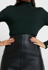 Dunkelgrüner, taillierter Rollkragenpullover, kombiniert mit einem schwarz-hochgeschnittenen Lederrock mit seitlichen Taschen und glänzender Textur.