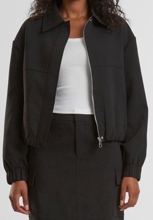 Kvinde iført en sort lynlåsflyver med elastiske manchetter over en hvid ribbet tanktop og matchende sorte bukser med sidelommer.