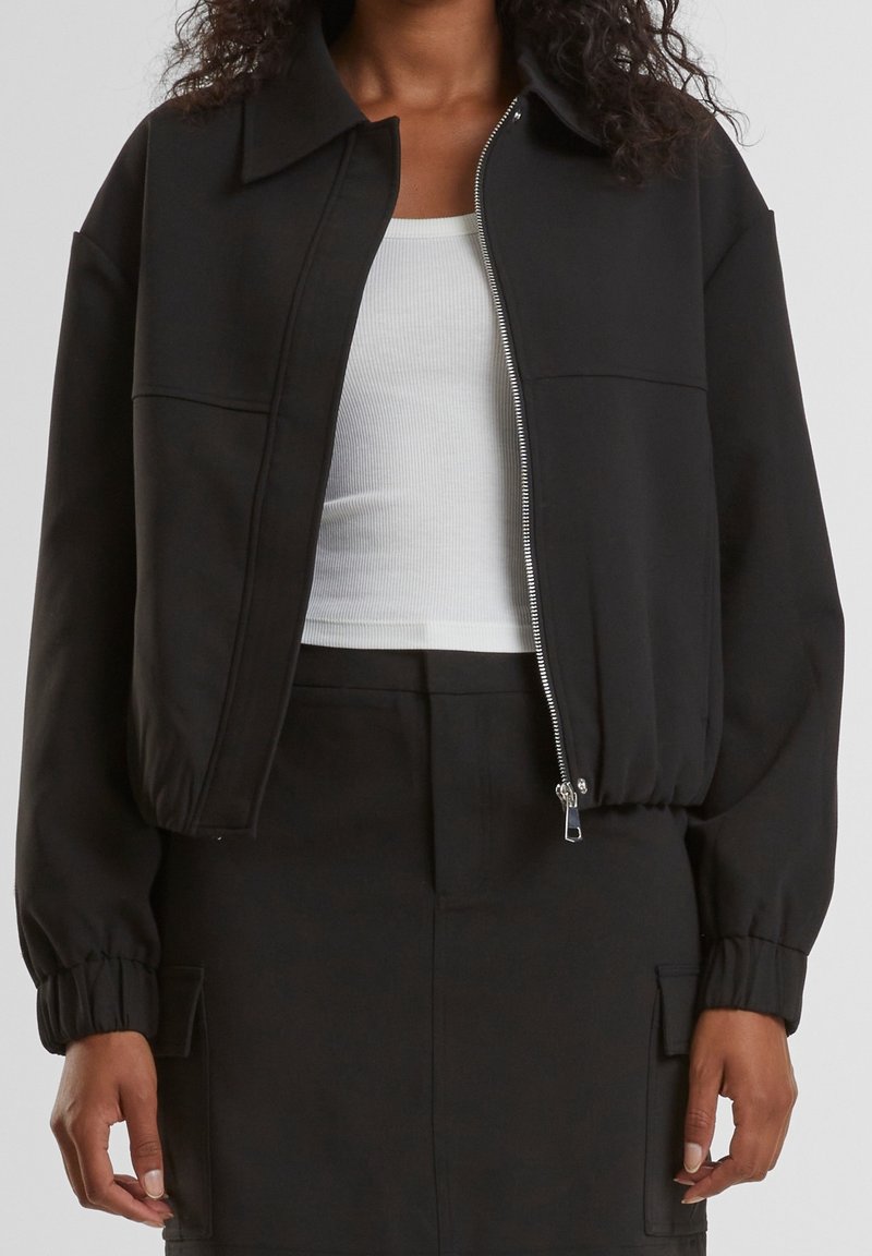 Femme portant une veste noire zippée avec des poignets élastiques sur un débardeur blanc côtelé et un pantalon noir assorti avec des poches latérales.