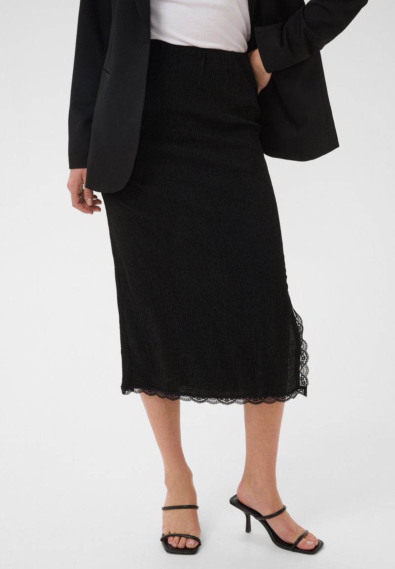 Femme portant une jupe midi noire avec bordure en dentelle, un blazer noir, un haut blanc et des sandales à talons noires sur fond blanc.