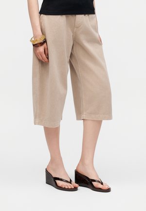 Beige vidde cropped bukser kombineret med sorte kile-sandaler og to gennemsigtige armbånd på venstre håndled, båret med en sort top.
