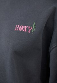 Svart hettegenser med myk tekstur, med en rosa og grønn "Roxy" logo på forsiden. Avslappet design med romslig passform.