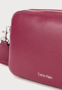 Bolso bandolera de Calvin Klein en cuero texturizado de color rosa oscuro con cremallera y herrajes plateados. Presenta una sutil marca en la parte frontal.