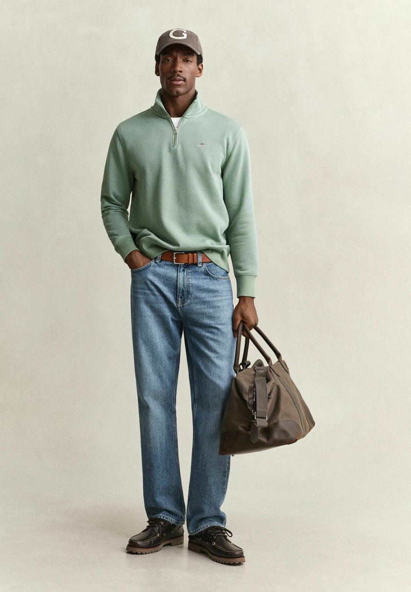 Man wearing green quarter-zip sweater, blue jeans, brown belt, cap, and boots holding a brown duffel bag against a plain background.