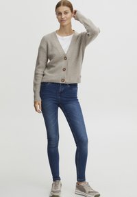 Beige stripete cardigan med treknapper, iført over en hvit T-skjorte og kombinert med blå skinny jeans og grå joggesko.