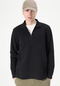 Schwarzer Pullover aus einem weichen Stoff, mit einem halben Reißverschluss-Kragen und langen Ärmeln. Getragen über einem hellen T-Shirt.