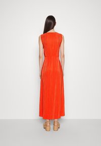 Langes, ärmelloses Kleid aus leuchtend orangefarbenem plissiertem Stoff. Enganliegende Taille mit einem fließenden Rock. Das Model trägt sandfarbene Plateausandalen.