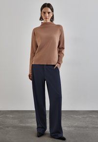 Brun stickad tröja med hög halskrage i kombination med marinblå vida byxor och svarta spetsiga skor mot en neutral bakgrund.