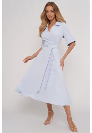 Kobieta w jasnoniebieskiej sukience kopertowej z podwiniętymi rękawami do połowy przedramienia i wiązanym paskiem w talii, pozująca w cielistych sandałach na wysokim obcasie na jednolitym tle.