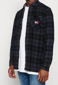 Uomo che indossa una camicia a quadri nera e blu scuro sbottonata sopra una maglietta bianca e jeans blu, con il logo "Tommy Jeans" sulla tasca del petto.
