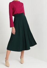 Gonna midi plissettata verde con cintura alta, abbinata a un top fucsia a maniche lunghe. Indossata con scarpe slip-on beige, su uno sfondo neutro.