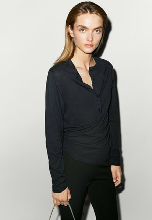 Giovane donna con capelli castano chiaro che indossa una camicia nera a maniche lunghe con bottoni e pantaloni neri, che tiene con la mano sinistra la maniglia di una borsa grigia.