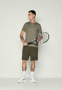 Olivu zaļas sporta T-krekls un šorti, ar vārdu "BORO" melnā krāsā uz šortiem. Modelis tur tenisa raketi, ģērbies baltos sporta apavos.