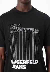 Svart bomull T-skjorte med dristig hvitt tekstdesign. Teksten inkluderer "KARL LAGERFELD" og "LAGERFELD JEANS" med vertikale linjer.