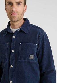 Giacca camicia in corduroy blu navy con due tasche sul petto, bottoni a pressione argentati e una toppa con il logo sulla tasca sinistra.