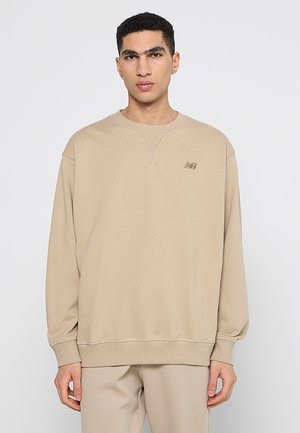 Junger Mann mit kurzen lockigen Haaren, trägt beige New Balance Sweatshirt und passende Hose, steht vor einem schlichten hellgrauen Hintergrund.