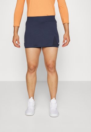 Jupe de sport marine avec une taille haute et une fente sur le côté, fabriquée en tissu extensible ; portée avec des baskets blanches pour un look sportif.