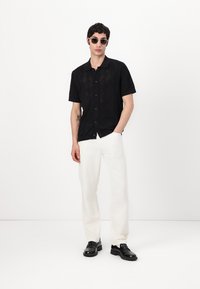 Camisa negra de manga corta con un patrón de diamantes texturizado, combinada con pantalones sueltos de color claro y zapatos negros. Lleva gafas de sol redondas.