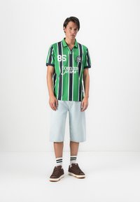 Young man wearing green and navy striped polo shirt, light denim knee-length shorts, white socks with black stripes, and brown sneakers.