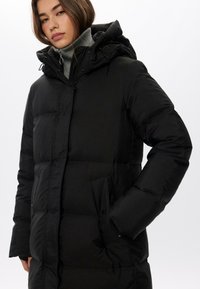 Černá péřová bunda s kapucí, s předním zipem, dvěma bočními kapsami a prošívanou texturou. Obsahuje vysoký límec a nastavitelné manžety.