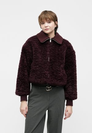 Ung person med kort hår iført en mørk lilla blød zip-up jakke og grå bukser med en hjerteformet bæltespænde.