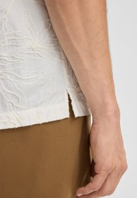 Witte geborduurde shirt met bloemenpatronen, met een licht gebogen zoom en een zichtbare naad aan de zijkant. Bruin stof zichtbaar eronder.
