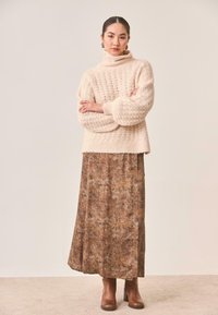 Suéter grueso de punto color crema con cuello alto, combinado con una falda larga marrón con efecto mármol y botas marrones con tacón.