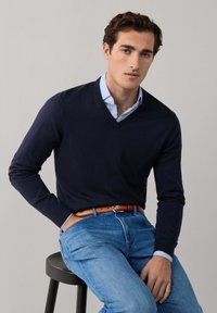 Giovane seduto su uno sgabello nero, indossa un maglione a V blu navy, una camicia a righe azzurre, jeans blu e una cintura marrone, su uno sfondo semplice.
