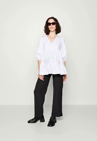 Blouse en coton blanc avec un col en V et un ourlet volanté, associée à un pantalon noir à larges jambes et des mocassins noirs. Le modèle porte des lunettes de soleil foncées.
