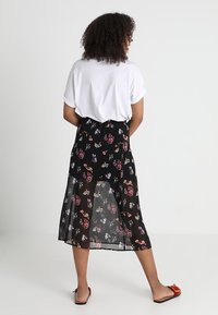 Nainen, jolla on kihara hius, pukeutuneena valkoiseen lyhythihaiseen paitaan, mustaan kukalliseen midi-hameeseen ja punaisiin liukukenkiin, seisoo valkoista taustaa vasten.