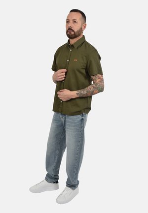 Uomo con braccio tatuato indossa una camicia verde oliva a maniche corte con bottoni, jeans azzurri e scarpe da ginnastica bianche, in piedi davanti a uno sfondo bianco.