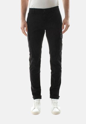 Uomo che indossa pantaloni cargo neri e scarpe da ginnastica bianche, in piedi contro uno sfondo bianco uniforme con le mani rilassate ai lati.