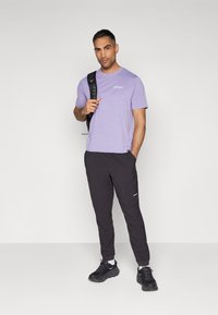 Paarse lichtgewicht t-shirt met geprinte logo, gecombineerd met zwarte taperende joggers en zwarte sportschoenen. Rugzak over één schouder geslingerd.
