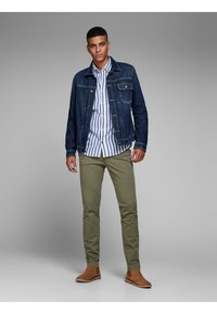 Chaqueta de mezclilla azul, camisa a rayas blanca y azul, pantalones verde olivo y botas de ante marrón. El modelo está de pie contra un fondo neutro.