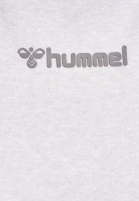 Logo de la marque de sport Hummel avec symbole d'abeille stylisé et nom de la marque en minuscules gris sur fond de tissu blanc.