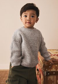 Maglione grigio lavorato a maglia con trama a costine, scollo rotondo e spalle scese, abbinato a pantaloni cargo verdi. Sfondo in legno naturale.