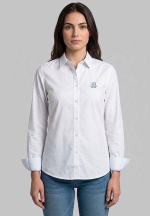 Mujer con una camisa blanca abotonada con un pequeño emblema azul en el pecho izquierdo, combinada con vaqueros azules, de pie contra un fondo liso.