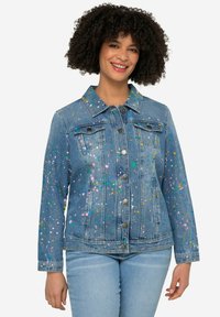 Veste en denim avec un délavage bleu clair, ornée de tâches de peinture multicolores, col classique, deux poches poitrine et fermeture par boutons.