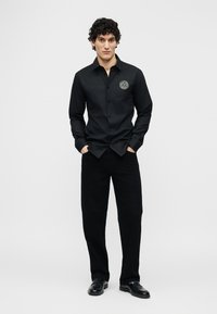 Άνδρας με σγουρά μαλλιά που φοράει μαύρο πουκάμισο με κουμπιά και λογότυπο στο στήθος, μαύρο παντελόνι και μαύρα παπούτσια, στέκεται με τα χέρια στις τσέπες.