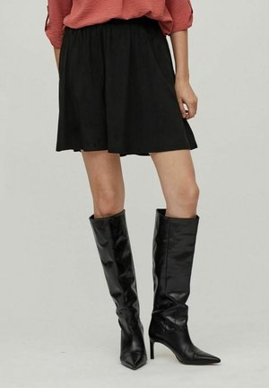 Person wearing a loose red top, black knee-length skirt, and tall black heeled boots, standing against a plain light background.