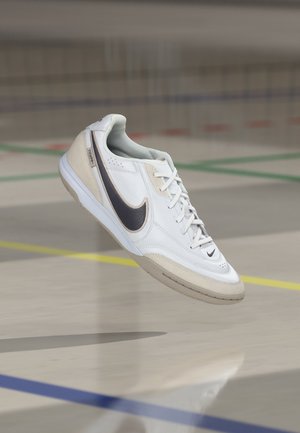 Λευκό και μπεζ αθλητικό παπούτσι Nike Tiempo με μαύρο σήμα να αιωρείται πάνω από τις σημάνσεις στο δάπεδο του εσωτερικού γυμναστηρίου.