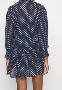 Kvinna i en långärmad klänning i marinblå med vita prickar, med en veckad volangkant och uppvikta ärmar, sedd bakifrån.