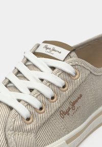 Baskets en toile beige avec des lacets blancs, présentant un motif rayé texturé, des œillets en métal et un patch logo sur la languette.