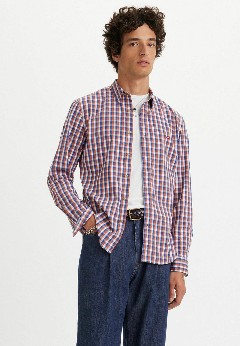Camisa a cuadros con patrones rojos, azules y blancos, de mangas largas, con cuello abotonado y un corte ajustado, combinada con jeans azul oscuro.