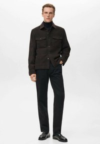 Chaqueta camisa marrón a cuadros con dos bolsillos en el pecho, cierre de botones y cuello, combinada con pantalones negros y zapatos negros. Textura suave.