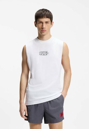 Młody mężczyzna ubrany w białą koszulkę bez rękawów z logo "HUGO" oraz ciemnoszare szorty z czerwoną naszywką z logo, stojący na białym tle.