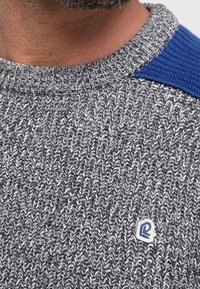 Pull tricoté gris et blanc avec un motif texturé, accents en tricot bleu sur l'épaule et petit badge logo à l'avant.