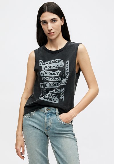 Young woman with long dark hair wears sleeveless black graphic tee and light blue studded jeans, standing with one hand in pocket.