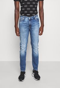 Blå denimjeans med slim fit, med en urtvättad design, fem fickor och dragkedja, kombinerat med en svart mönstrad t-shirt och svarta sneakers.