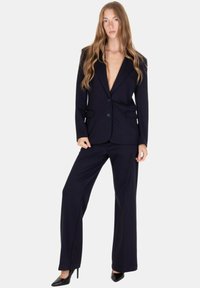 Completo gessato blu navy con blazer su misura dal profondo scollo a V, pantaloni abbinati a gamba larga e décolleté nere appuntite. Tessuto dalla texture liscia.